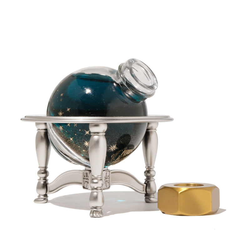 Ferris Wheel Press's 20ml Ink Carriage (Forged Edition) features a deep blue, star-flecked ink in a spherical glass bottle with a gold cap and silver pedestal.