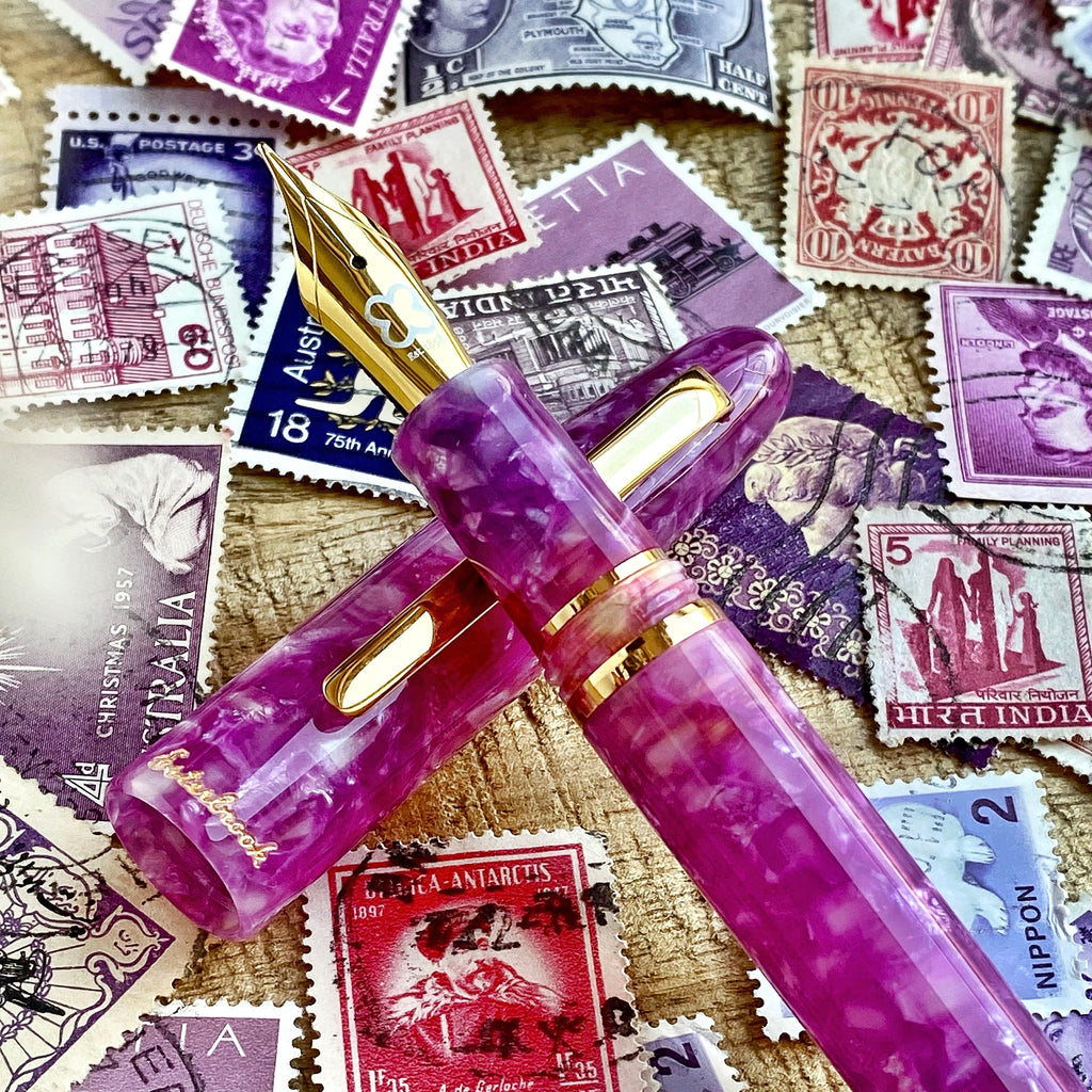 Esterbrook Estie - Punch / Gold - Techo Custom Nib fountain pen with gold trim, resting on vintage stamps, showcasing elegant purple-marbled acrylic craftsmanship.
