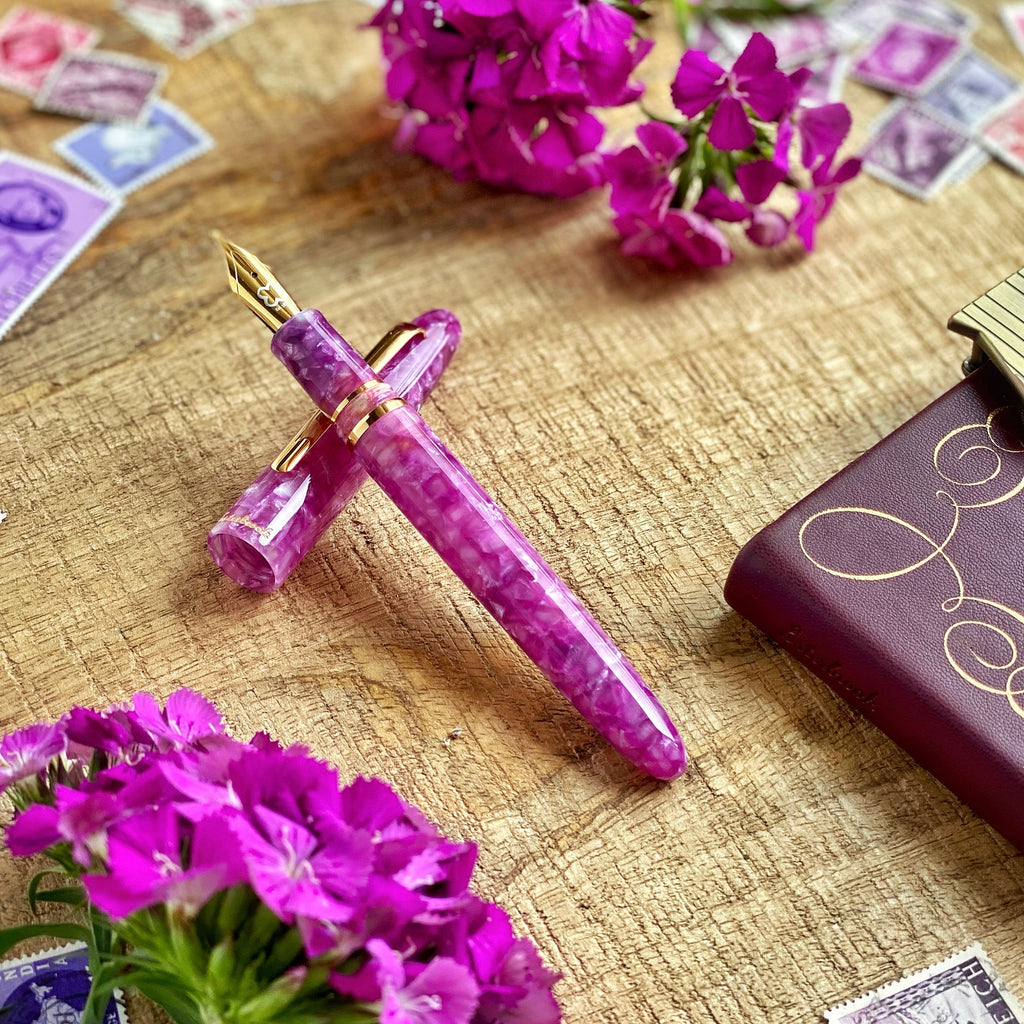 Esterbrook Estie - Punch / Gold - Techo Custom Nib fountain pen, marbled lavender-pink resin with gold trim, surrounded by magenta flowers and a burgundy leather notebook in a still-life setting.