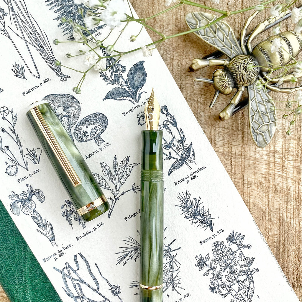 Esterbrook JR Pocket Pen - Palm Green - Journaling (Gena Custom) features a green acrylic body with gold trim, exposed nib, on botanical print background with desk accessories.