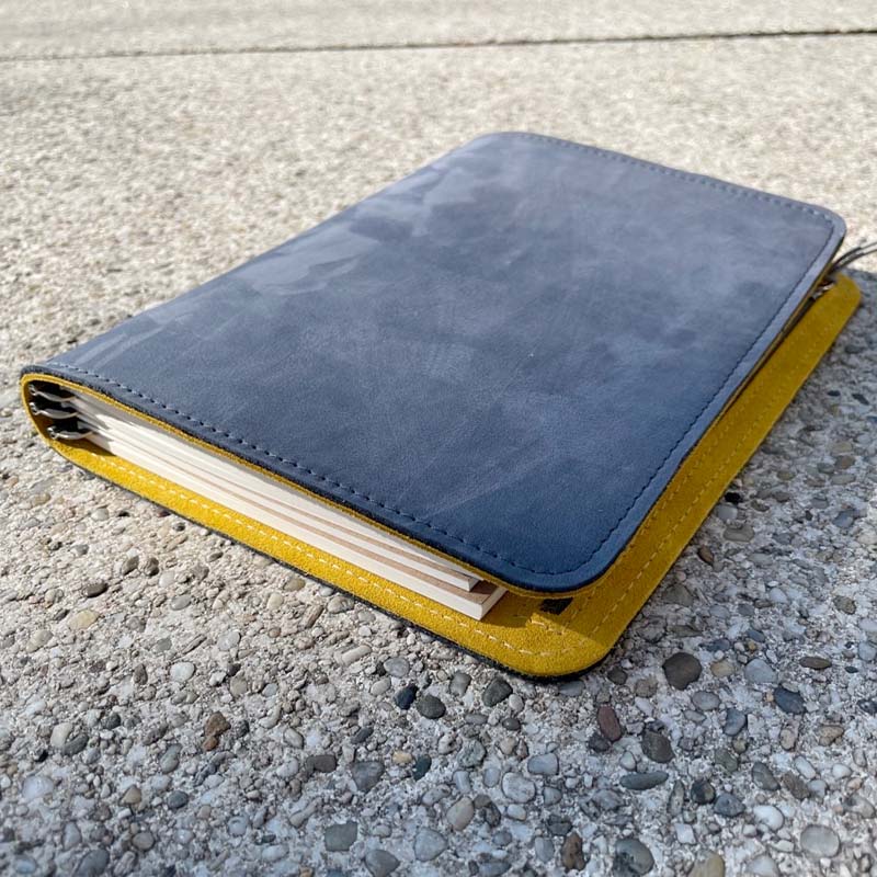 Roterfaden Taschenbegleiter SO_22 planner with suede-like smoke blue cover and mustard lining, closed on a concrete surface, showcasing its flexible, eco-friendly leather design.