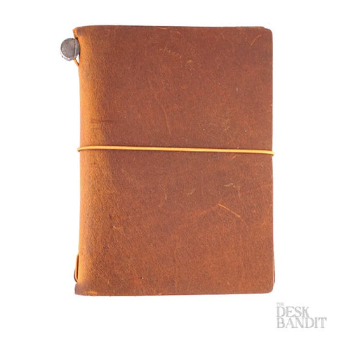 Passport Leather Cover (Camel) shown as a rustic-style, brown leather notebook with a single elastic band wrapping around its cover, highlighting its handmade quality and unique texture.