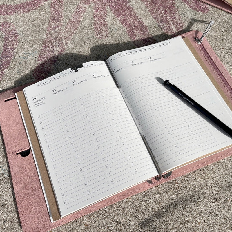 Taschenbegleiter SO_23 by Roterfaden, a weekly planner with a soft-pink cover, open inside with a black pen resting on blank pages.