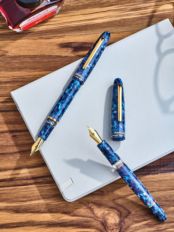 Esterbrook Estie Blue Moon fountain pens with gold trim and fine nib, displayed on a notebook with red fountain pen ink bottle, showcasing elegant design.