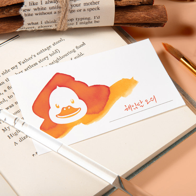 Wearingeul 'White Duck' Ink Swatch Cards, showcasing a cartoon duck illustration, rest on an open book alongside a glass dip-pen and paintbrush. Pack of 50, fountain pen friendly.