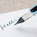 Teranishi Chemical Industry Glass Pen (Sunset Pink) writing Hello! on graph paper, showcasing its easy-to-clean glass nib and portable cap design.