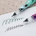 Teranishi Chemical Industry Glass Pen (Sunset Pink) with mint-green and purple handles on graph paper, showcasing green and purple ink strokes. Easy to clean and cap-protected.