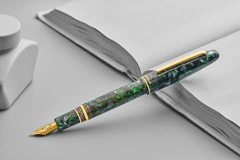 Esterbrook Estie - Jurassic / Gold - Fude fountain pen with green-blue marbled finish and gold trim, beside white ink bottle, on open notebook.