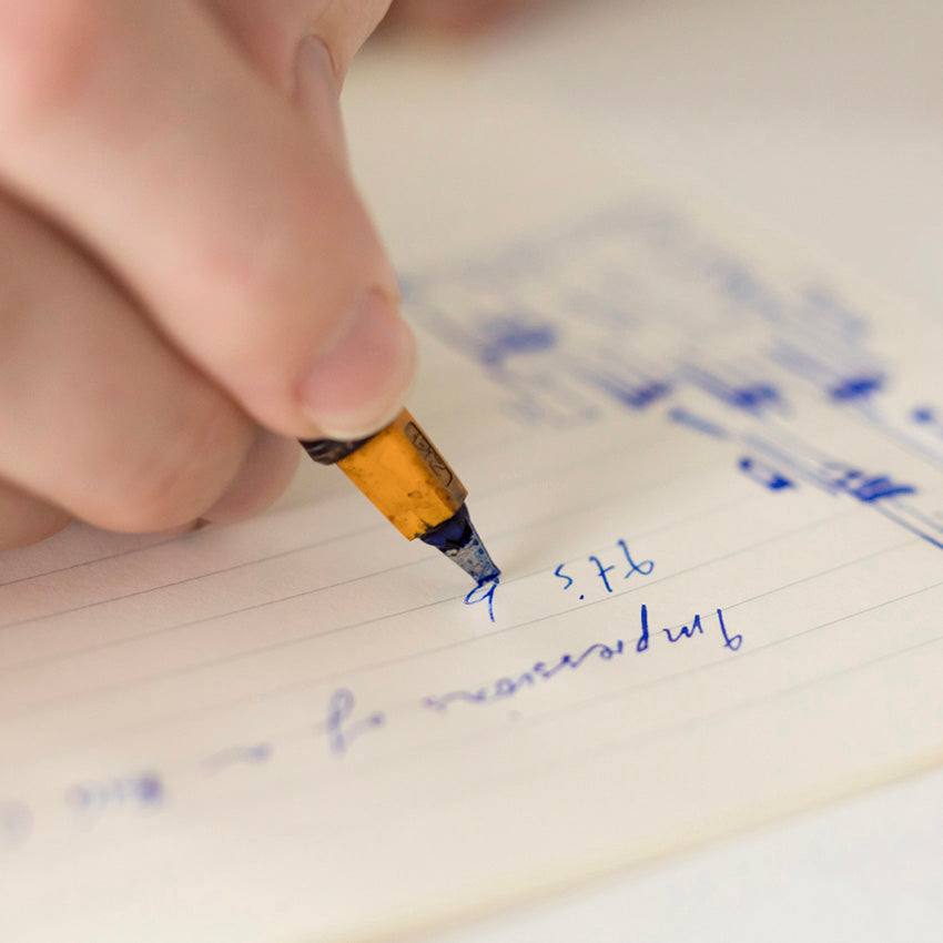 Hand using a fountain pen on Midori MD Notebook Light, B6 Slim Grid. Blue ink cursive writing and sketches visible on lined paper.