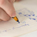 Hand using a fountain pen on Midori MD Notebook Light, B6 Slim Grid. Blue ink cursive writing and sketches visible on lined paper.