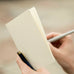 Hands holding a cream-colored MD Notebook Light – B6 Slim Grid, with a silver pen poised to write, and a blue pen clipped to the cover.