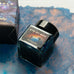 Journalize Eye of God Shimmer fountain pen ink, 30ml, in a square glass bottle with black cap, displayed with matching packaging on a desk surface.