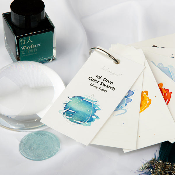 Wearingeul Ink Drop Swatch Cards (Ring Type) with colorful ink splashes, alongside Wayfarer teal fountain pen ink, magnifier, and decorative quill on a desk setup.