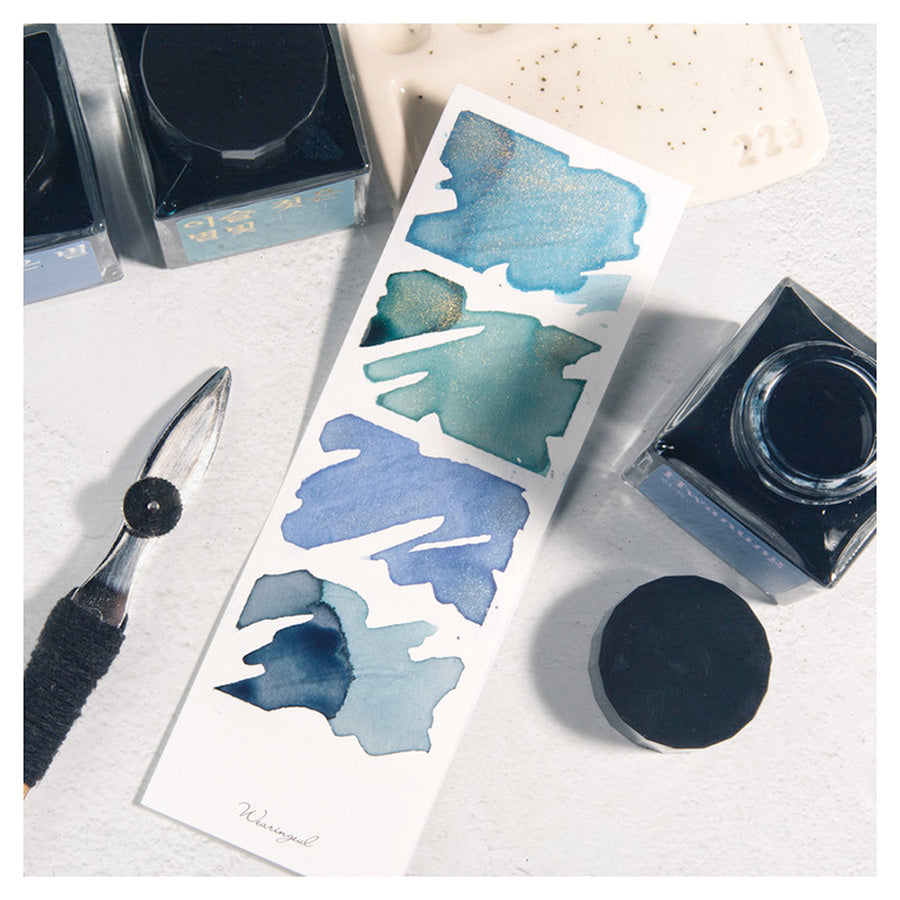 Wearingeul Four Photos Color Swatch (30 Sheets, White) with blue-green ink blobs, surrounded by ink bottles, ceramic palette, and nib tool. Ideal for fountain pen ink swatching.