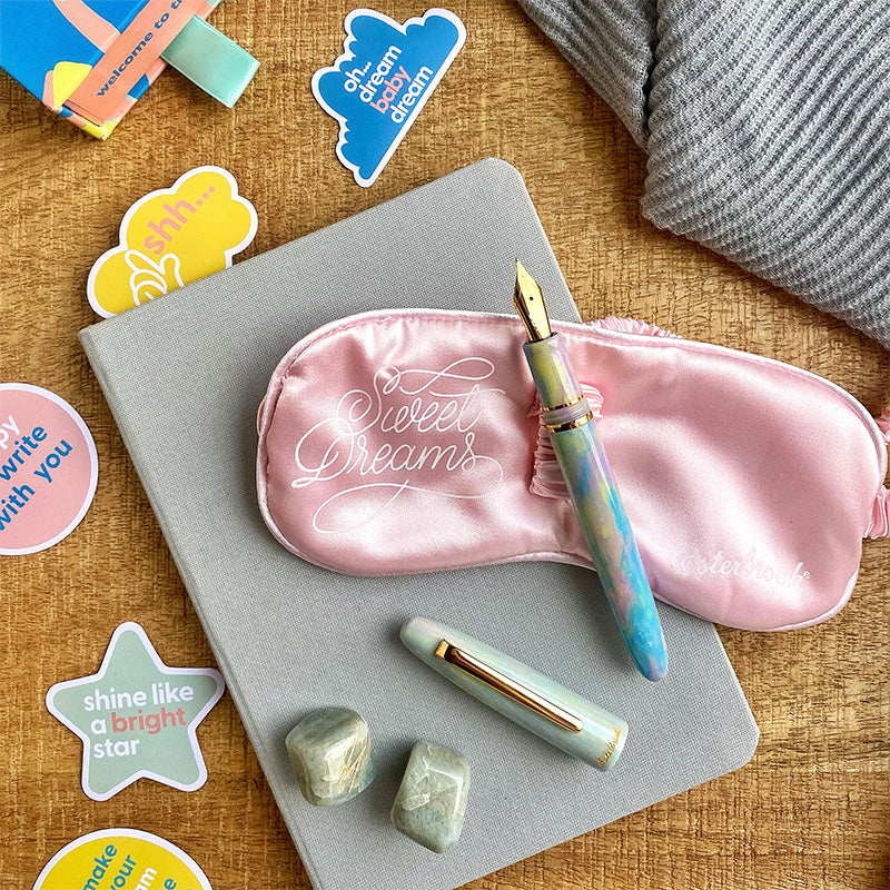 Esterbrook Estie - Sweet Dreams fountain pen with gold trims, journaling nib, alongside a pink satin sleep mask, light gray notebook, and stickers, on a wooden surface.