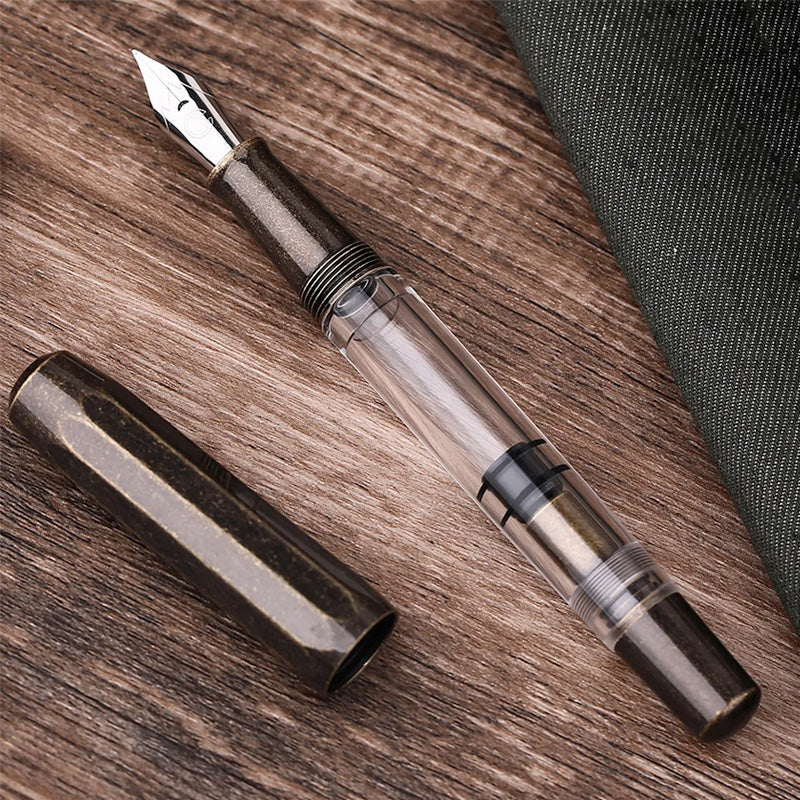 Moonman/Majohn T1 (Brass Version) fountain pen with transparent barrel, revealing piston mechanism, and bronze-speckled cap, fine steel nib, on wooden surface.