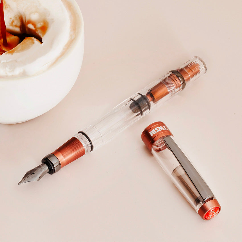 Diamond 580 ALR (Affogato with Onyx) - Stub 1.1 fountain pen, showcasing its clear body and orange-red metal trim, alongside a coffee mug with whipped cream.