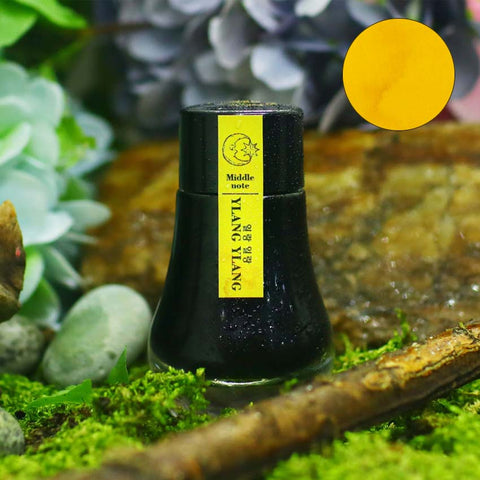 Dominant Industry Ylang Ylang (Scented) fountain pen ink, 25ml, in a dark glass bottle with a yellow label, displayed on moss with natural elements.