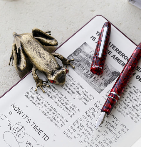 Esterbrook Tree Frog Page Clip, a brass-colored desk accessory, with a red-and-black marbled fountain pen and an open brochure, symbolizing resilience and creativity from Desk Bandit.