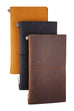 Three leather-bound notebooks stand upright, featuring a distressed brown Leather Cover, secured by elastic bands with metal studs, hand-made for Traveler's Company.