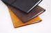 Three Traveler's Company leather covers stacked, featuring the Leather Cover (Camel) beneath, with elastic band closure, showcasing hand-crafted quality for notebook protection.