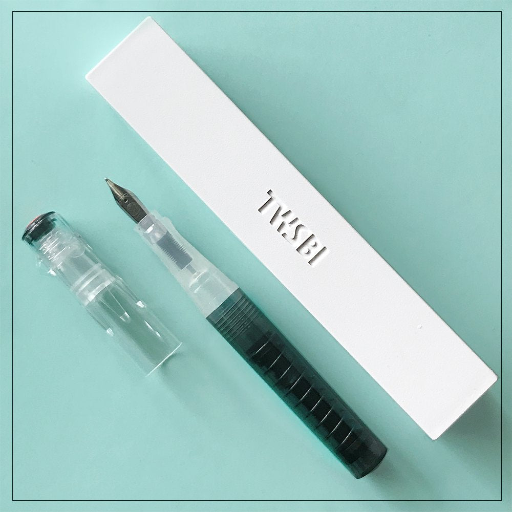 TWSBI GO (Smoke) - Extra Fine fountain pen, transparent with green ink reservoir, beside its embossed white TWSBI box on a teal surface.