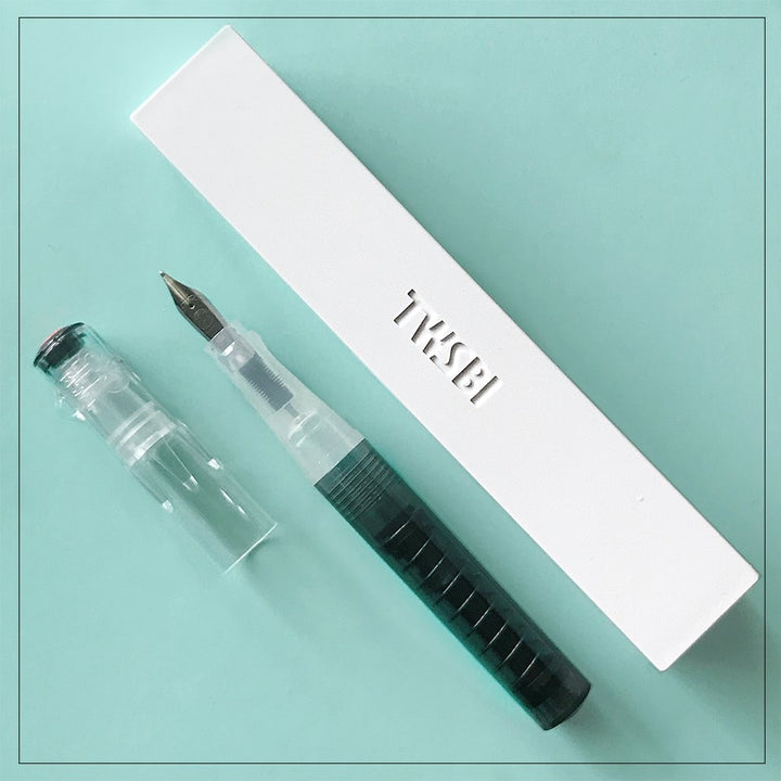 TWSBI GO (Smoke) - Extra Fine fountain pen, transparent with green ink reservoir, beside its embossed white TWSBI box on a teal surface.