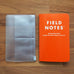 Zipper Case/Card Holder- Pocket on left, clear plastic with card slots and zippered pouch, next to an orange FIELD NOTES memo book on a wood surface.