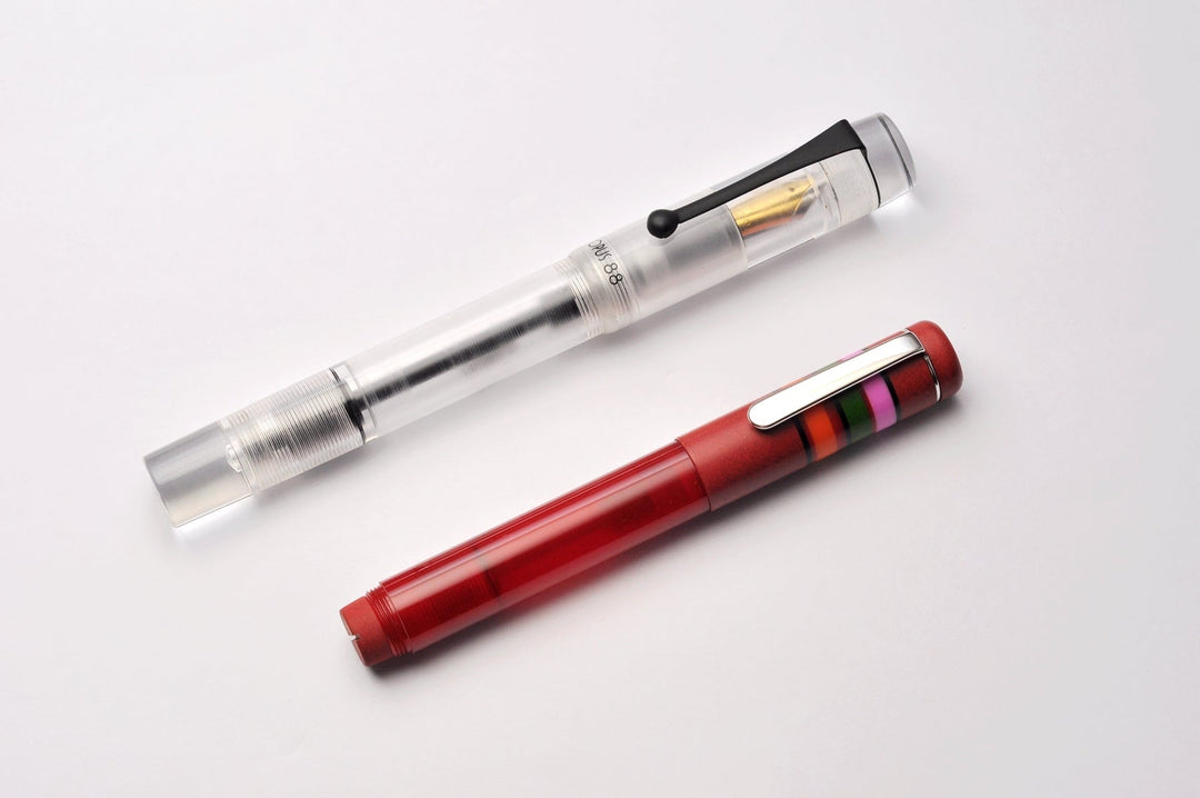 Two fountain pens; a transparent demonstrator with gold nib and black clip, alongside the OPUS 88 Fantasia - Red (Medium) with silver clip and colored band.